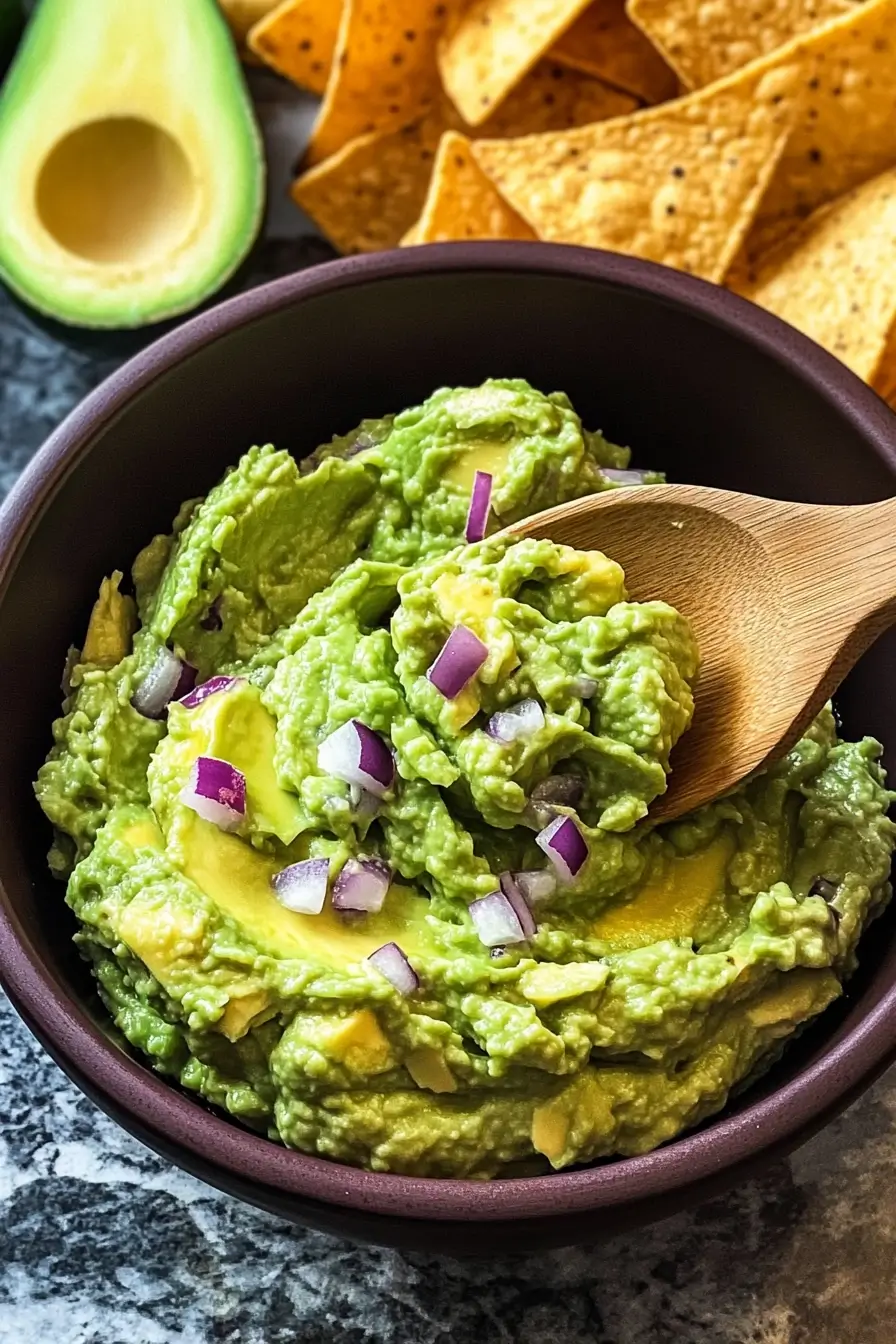 A bowl of creamy guacamole garnished with chopped red onions, with a wooden spoon scooping a portion, surrounded by tortilla chips and a halved avocado in the background.