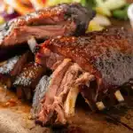 Glazed barbecue ribs with a side of sauce and fresh vegetables on a rustic wooden board.