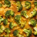 The image shows a delicious-looking dish of broccoli baked with a creamy, cheesy sauce. The broccoli florets are well-coated with the sauce and appear tender yet vibrant green, suggesting they have been cooked to maintain some crispness. Some florets are sprinkled with golden breadcrumbs, adding a crunchy texture contrast to the creamy sauce beneath. The overall appearance is appetizing and suggests a comfort food that integrates healthy ingredients.