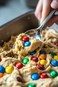 A close-up shot of peanut butter M&M cookie dough being scooped with a spoon, showing colorful M&Ms embedded in the creamy dough.