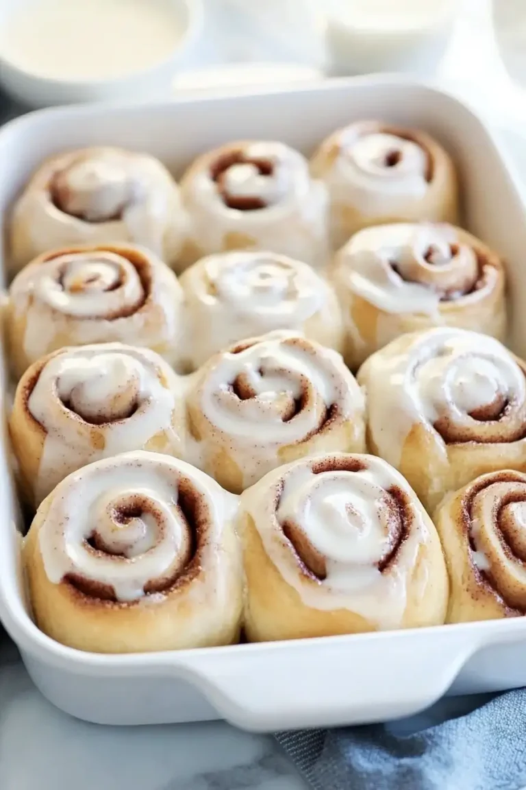 Freshly baked cinnamon rolls with creamy white icing, arranged in a white baking dish.
