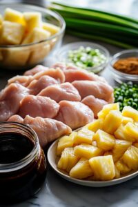 Ingredients for Sweet Hawaiian Chicken, including raw chicken, pineapple chunks, soy sauce, green onions, spices, and other fresh ingredients on a countertop.