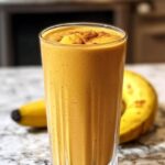 A tall glass of creamy Pumpkin Protein Smoothie garnished with a sprinkle of cinnamon, placed on a marble countertop with a ripe banana in the background.