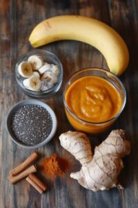  Ingredients for a Pumpkin Protein Smoothie, including a banana, pumpkin puree, chia seeds, ginger root, cinnamon sticks, and turmeric, arranged on a wooden surface.