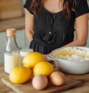 A woman in a black dress stands behind a table with ingredients for Fluffy Lemon Ricotta Pancakes, including lemons, eggs, milk, and flour.