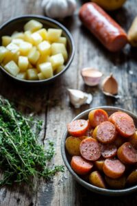  Ingredients for Potato Kielbasa Soup on a rustic wooden surface, including bowls of diced potatoes, sliced kielbasa, fresh thyme, garlic cloves, and sausage.