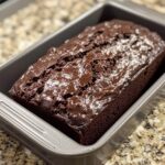 A freshly baked loaf of Double Chocolate Zucchini Bread in a metal loaf pan, with a glossy, chocolatey crust resting on a granite countertop.