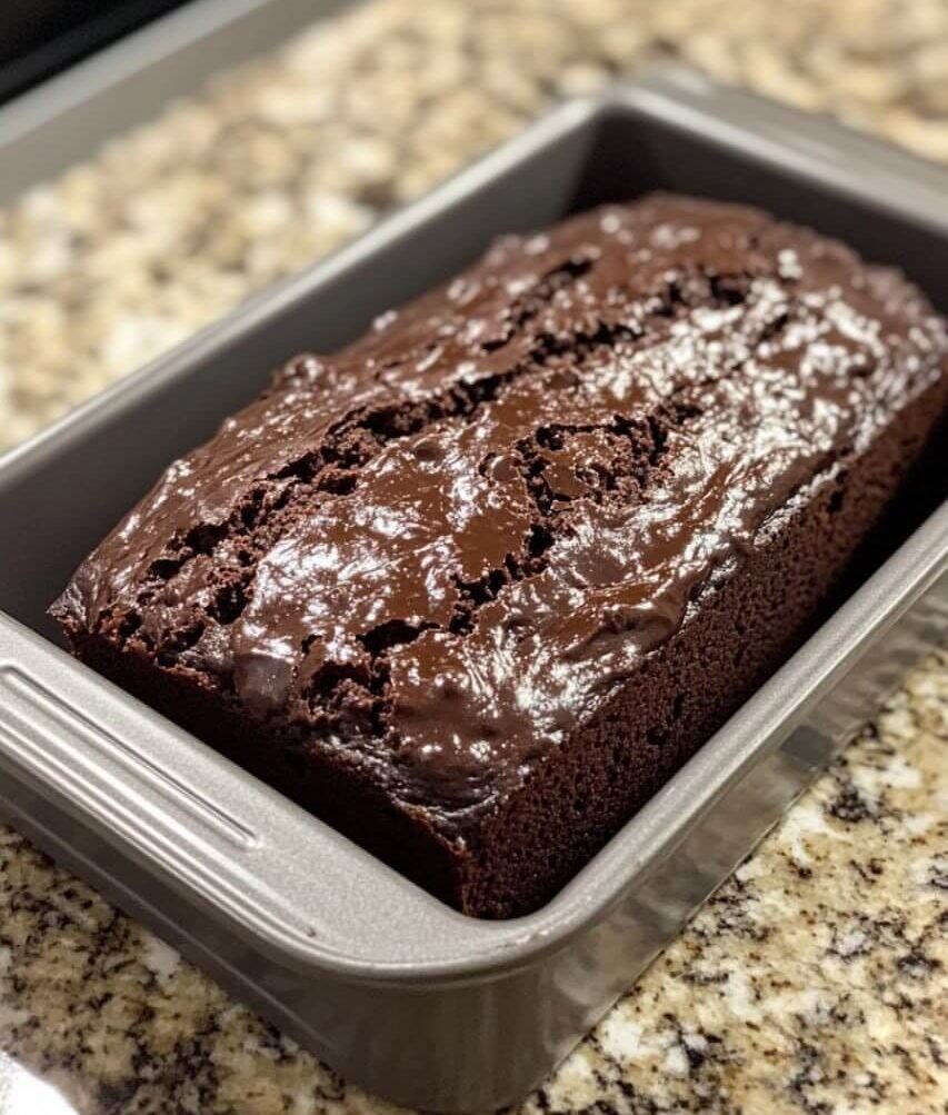 A freshly baked loaf of Double Chocolate Zucchini Bread in a metal loaf pan, with a glossy, chocolatey crust resting on a granite countertop.