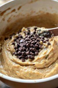 A mixing bowl filled with batter for Double Chocolate Zucchini Bread, with chocolate chips on top and a wooden spoon partially submerged.