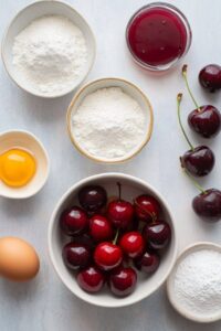 Ingredients for cherry pie bars, including fresh cherries, flour, sugar, eggs, and cherry filling, arranged on a light background