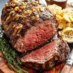 Perfectly cooked smoked garlic butter rib roast sliced on a wooden board, garnished with fresh thyme and served with roasted potatoes and a side of sauce.