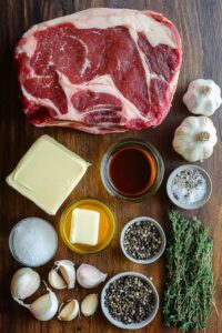 Ingredients for smoked garlic butter rib, including a raw rib cut, garlic, butter, thyme, pepper, salt, and marinade, arranged on a wooden surface.