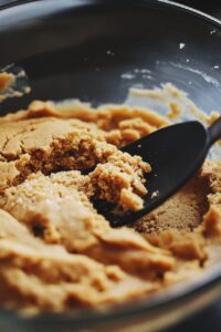  A close-up of peanut butter cookie dough being mixed in a bowl with a black spatula.Title: Peanut Butter Cookie Dough Preparation