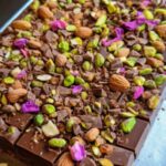 A slab of Dubai-inspired chocolate bar topped with pistachios, almonds, rose petals, and chocolate chunks on a tray.