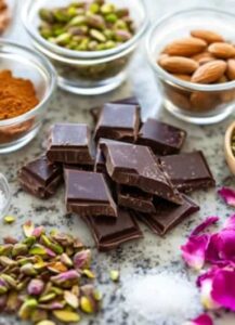 Ingredients for a Dubai-inspired chocolate bar, including dark chocolate pieces, pistachios, almonds, rose petals, and cocoa powder, arranged on a marble countertop.