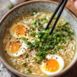 A bowl of egg drop soup with soft-boiled eggs, chopped green onions, and seasonings, with chopsticks picking up noodles.