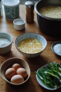  Ingredients and a bowl of egg drop soup arranged on a wooden table, including eggs, green onions, and seasonings.