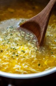 A pot of freshly made egg drop soup with herbs, stirred with a wooden spoon. Title: Freshly Prepared Egg Drop Soup