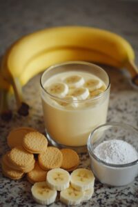 A glass of banana pudding topped with banana slices, surrounded by fresh bananas, vanilla cookies, and a small bowl of powdered sugar.