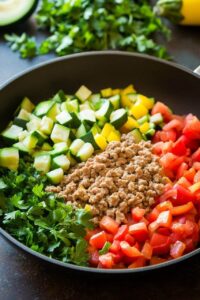 A skillet filled with fresh ingredients including ground turkey, chopped zucchini, yellow bell peppers, tomatoes, and parsley.