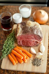 A raw chuck roast surrounded by fresh carrots, garlic cloves, thyme sprigs, black pepper, and seasoning ingredients on a wooden cutting board.