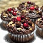 Chocolate cranberry oat muffins topped with fresh cranberries, chocolate chips, and oats, served on a white plate.