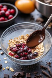 A mixing bowl with Chocolate Cranberry Oat Muffins, dried cranberries, and a spoon drizzling melted chocolate, surrounded by ingredients for baking.