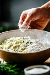 A hand mixing flour with shredded zucchini in a bowl, preparing the dough for zucchini bread.