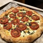 A freshly baked Tomato Ricotta Galette with golden puff pastry, creamy ricotta filling, and vibrant tomato slices garnished with fresh herbs.