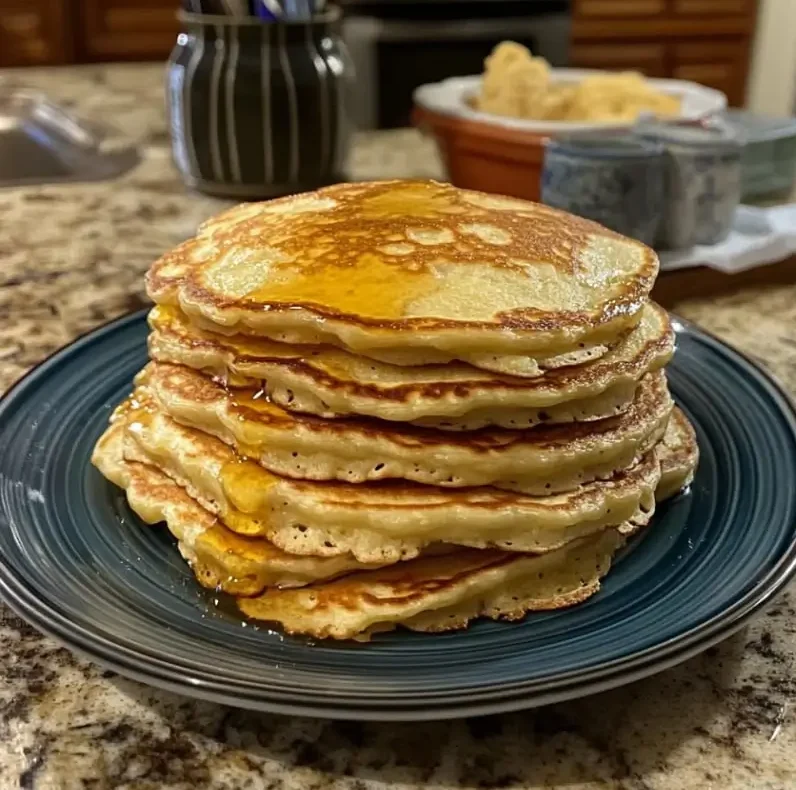 A stack of homemade pancakes drizzled with syrup, served on a dark plate, with a kitchen setup in the background