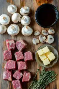 Raw ingredients for garlic butter steak bites with mushrooms, including cubed steak, whole and sliced mushrooms, butter cubes, fresh thyme, and a bowl of sauce, arranged on a wooden cutting board.