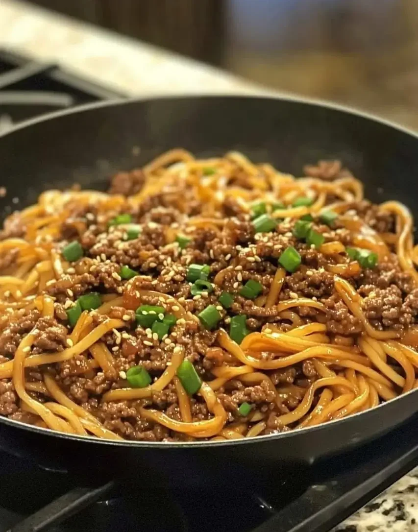 A skillet filled with Mongolian ground beef noodles, coated in a savory sauce, garnished with sesame seeds and fresh green onions.