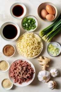 Fresh ingredients for Mongolian ground beef noodles, including ground beef, ramen noodles, soy sauce, eggs, green onions, garlic, ginger, and sesame seeds, arranged on a white surface.