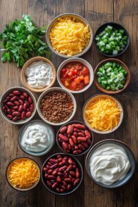 An assortment of chili ingredients, including shredded cheddar cheese, kidney beans, ground beef, sour cream, diced green onions, salsa, and fresh cilantro, arranged on a rustic wooden table.