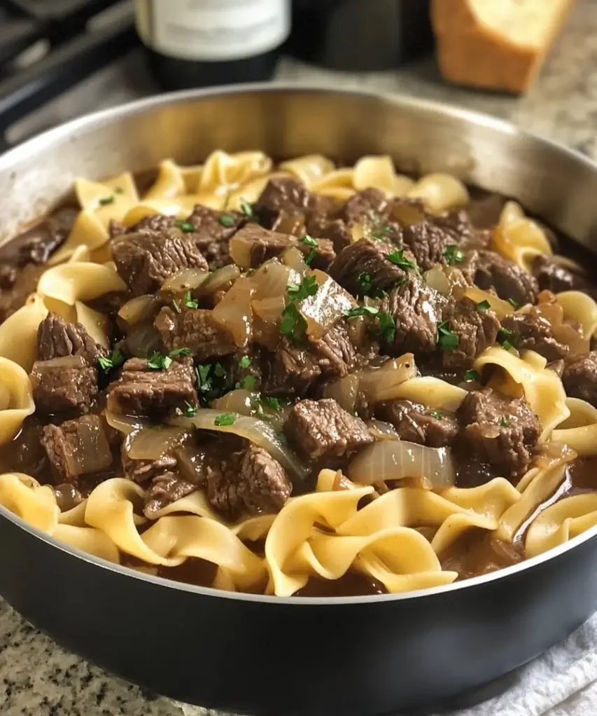 A skillet filled with French Onion Beef and Noodles, featuring tender beef chunks, caramelized onions, and egg noodles in a rich, savory broth, garnished with fresh parsley.