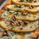 Close-up of a flatbread topped with sliced pears, crumbled blue cheese, chopped walnuts, and fresh herbs.